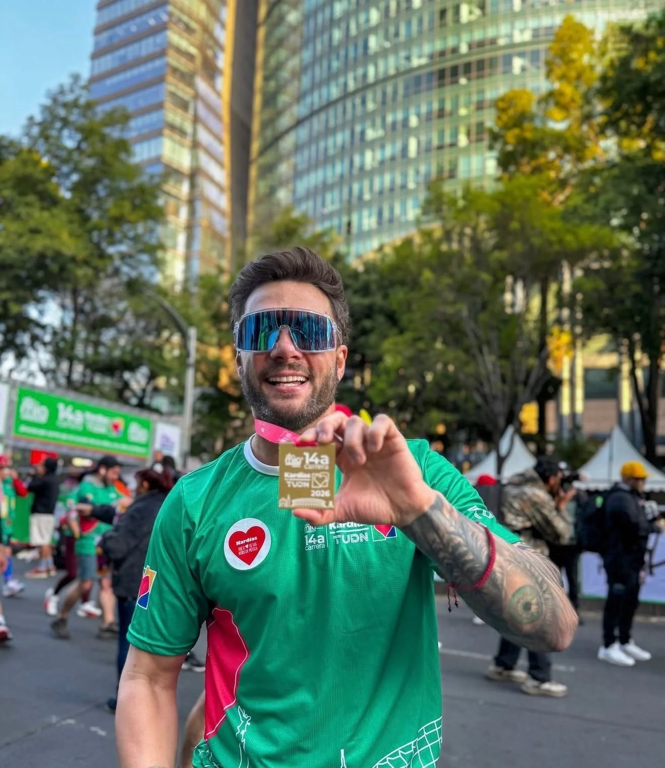 Un hombre sonriente en una carrera, sosteniendo una medalla en una mano, lleva gafas de sol y camiseta verde, en un entorno urbano con edificios y árboles.