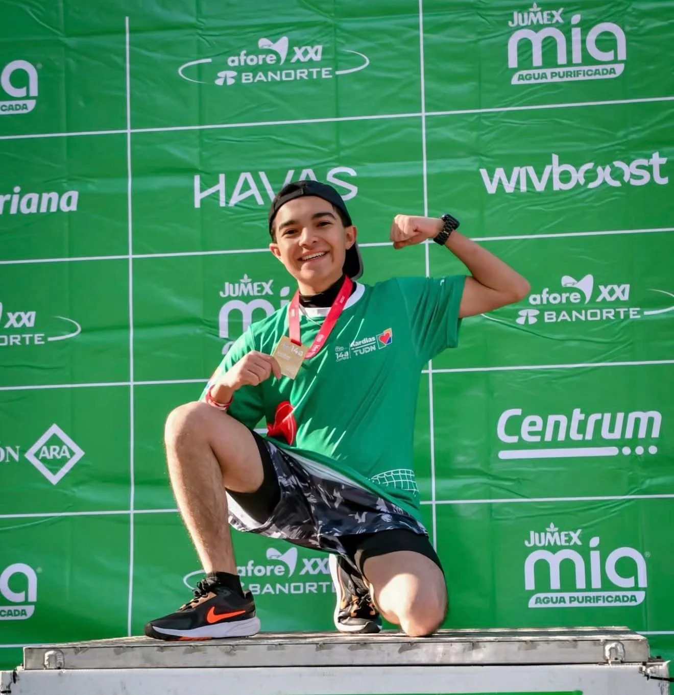 Joven deportista en attire deportivo, posando en una ceremonia de premiación, sosteniendo una medalla y haciendo un gesto de fuerza con el brazo, en un fondo verde con logotipos patrocinadores.