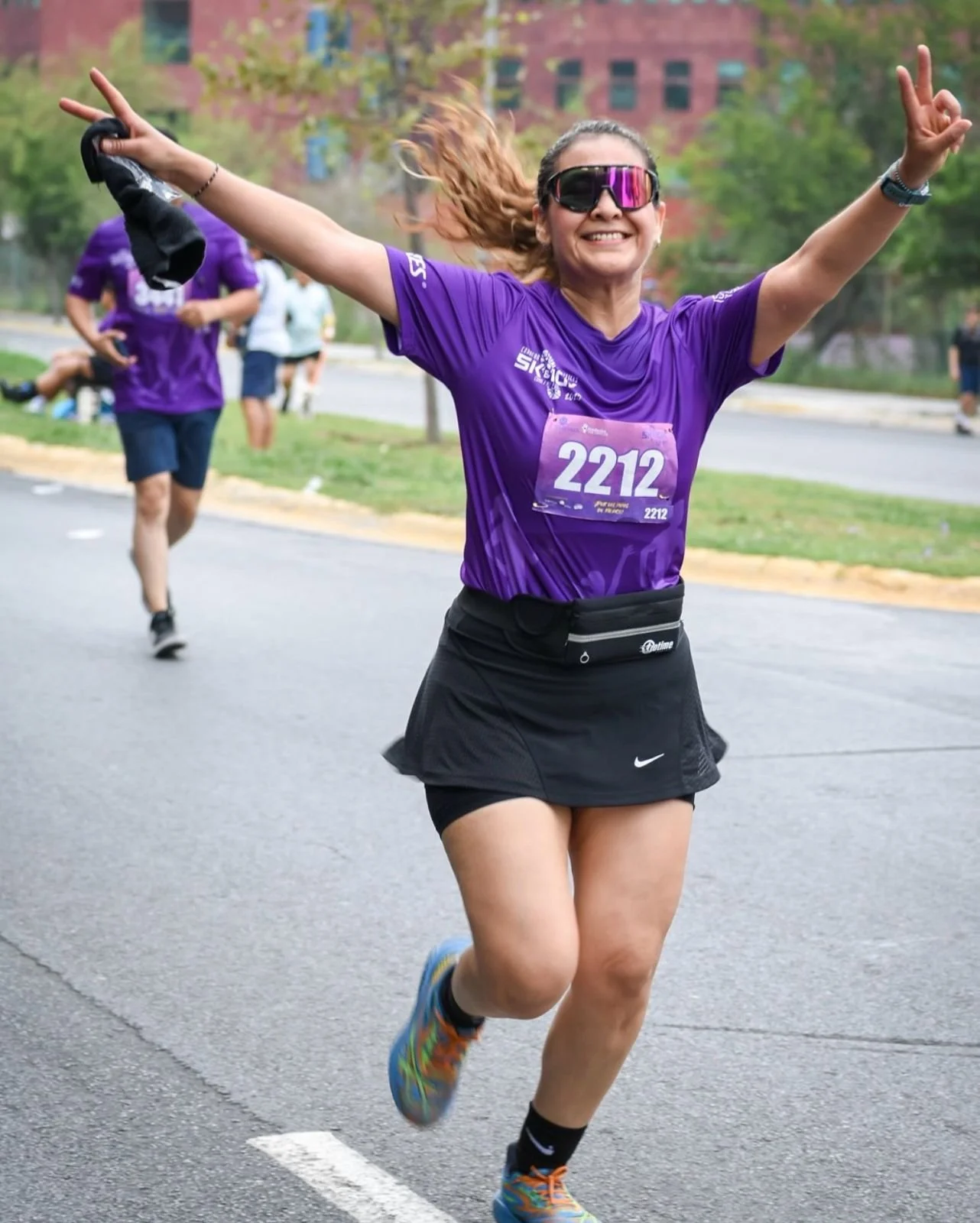 Una mujer sonriente y con gafas de sol corre en una carrera con camiseta morada y número 2212, levantando los brazos en señal de celebración.