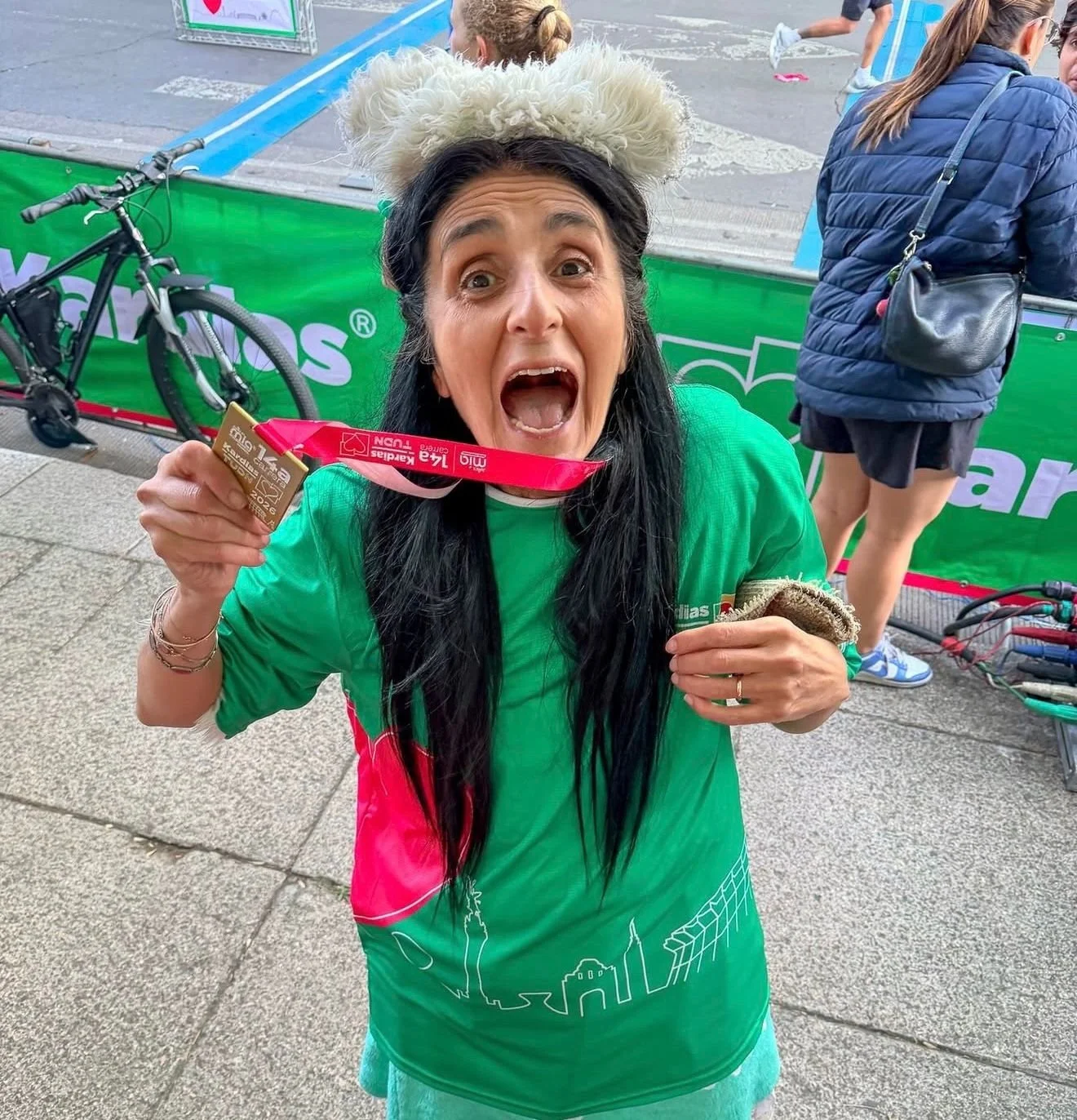 Mujer sorprendida y feliz que muestra una medalla de finisher, con orejas de oso de peluche en la cabeza, en medio de una carrera, con personas y bicicletas en el fondo.