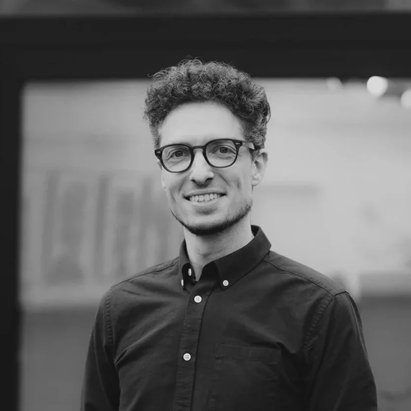 Photo en noir et blanc d'un homme souriant portant des lunettes et une chemise à boutons, dans un environnement extérieur