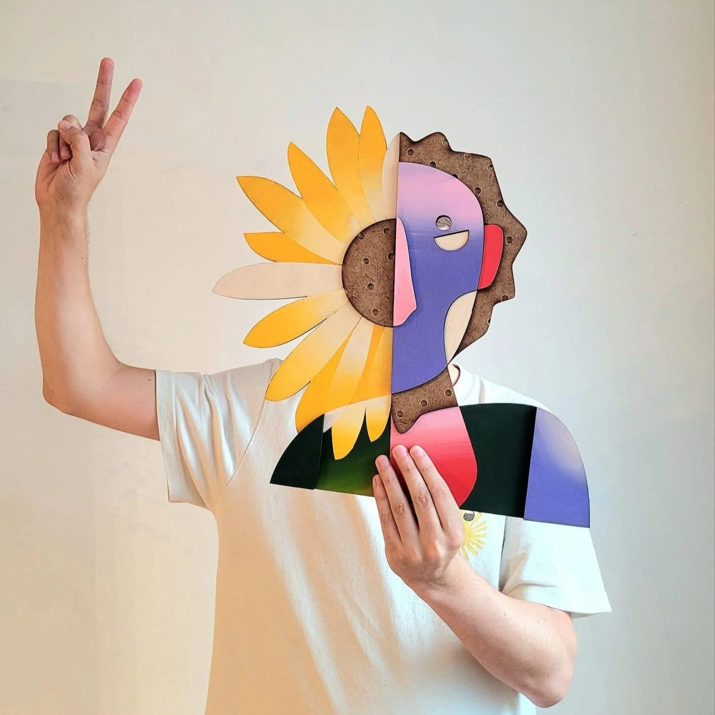 Person tenant un masque de carton représentant un visage stylisé, moitié soleil avec rayons jaunes, moitié visage humain avec une expression souriante, portant un t-shirt blanc.