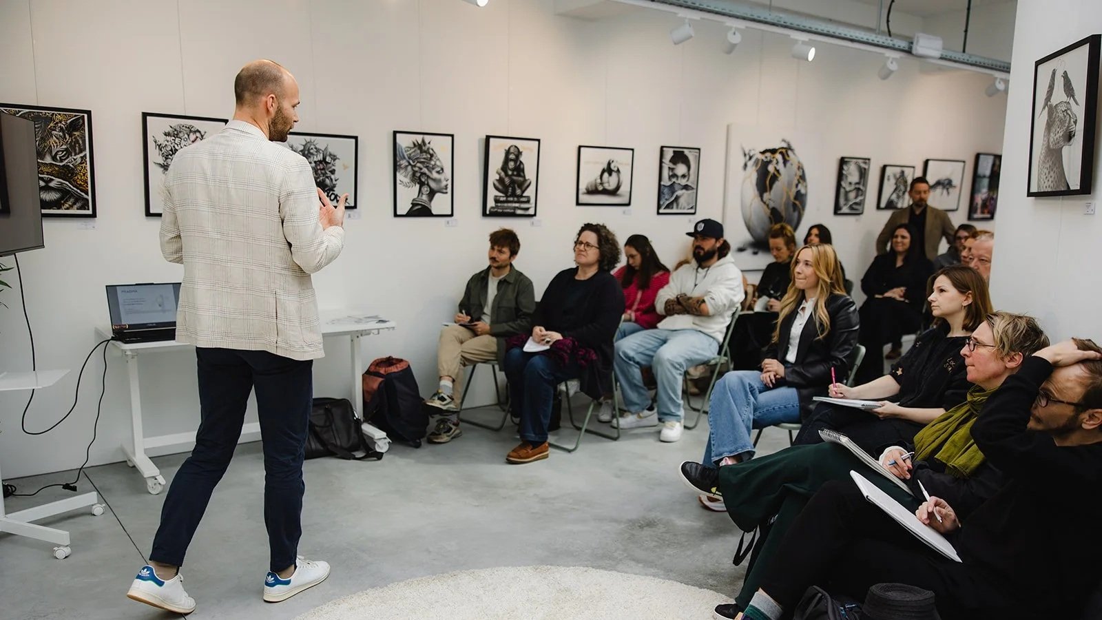 Un homme donne une présentation devant un groupe de personnes assises dans une galerie d'art.