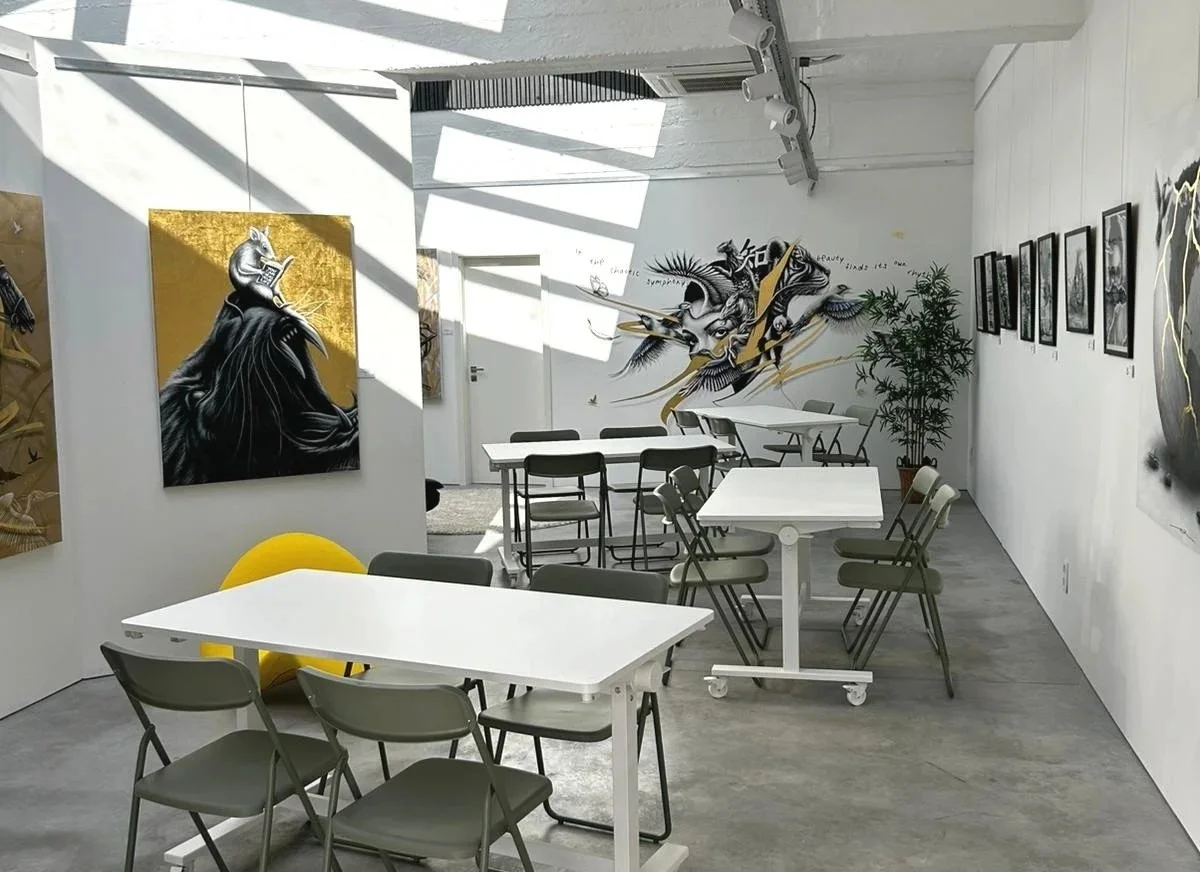 Une salle d'exposition d'art avec plusieurs peintures et dessins muraux, des tables et des chaises, un arbre en pot, et un éclairage suspendu.