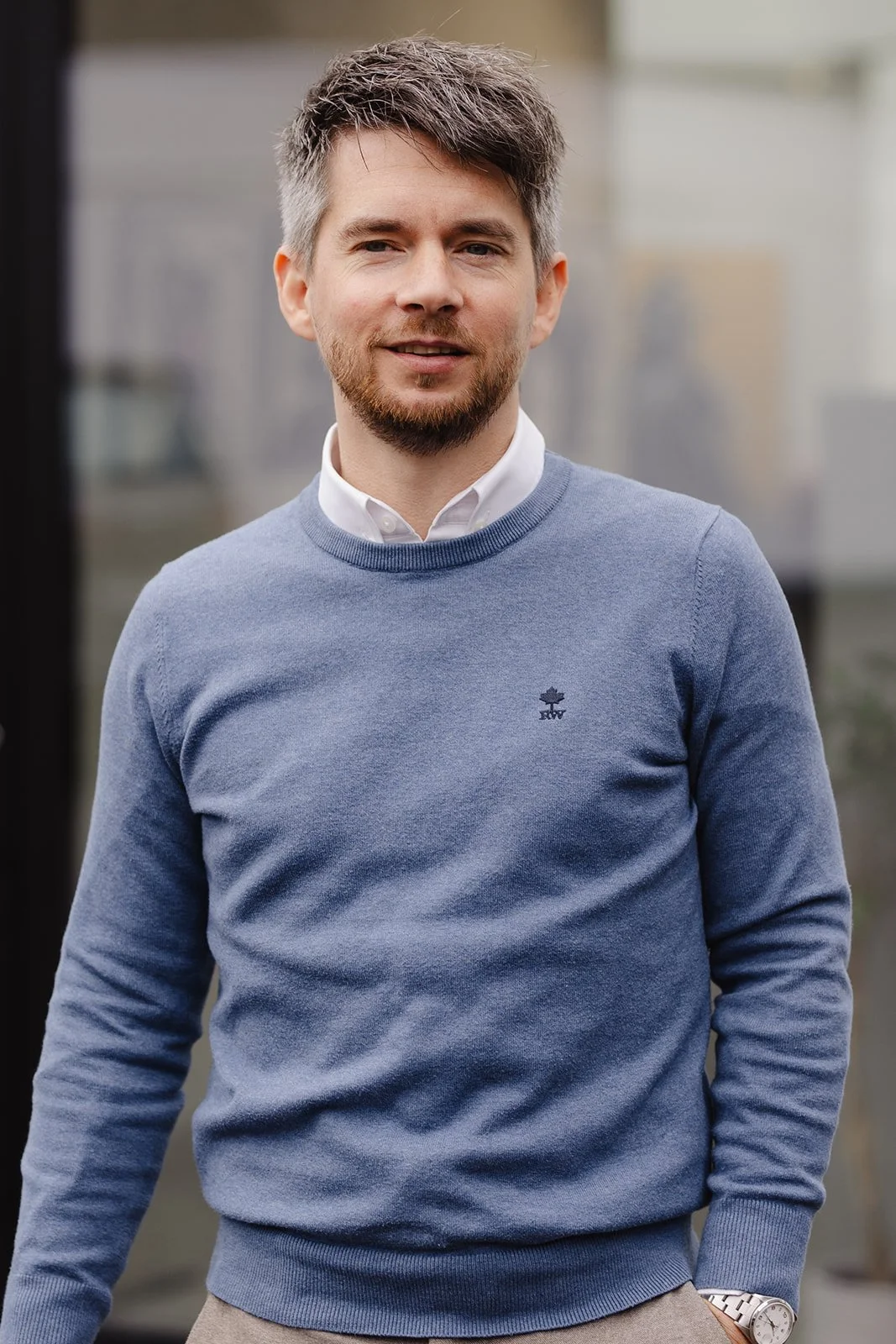 Un homme avec des cheveux bruns, une barbe légère, portant un pull bleu avec un logo d'une feuille d'érable, un col blanc, et un pantalon beige, posant à l'extérieur devant un bâtiment en verre.