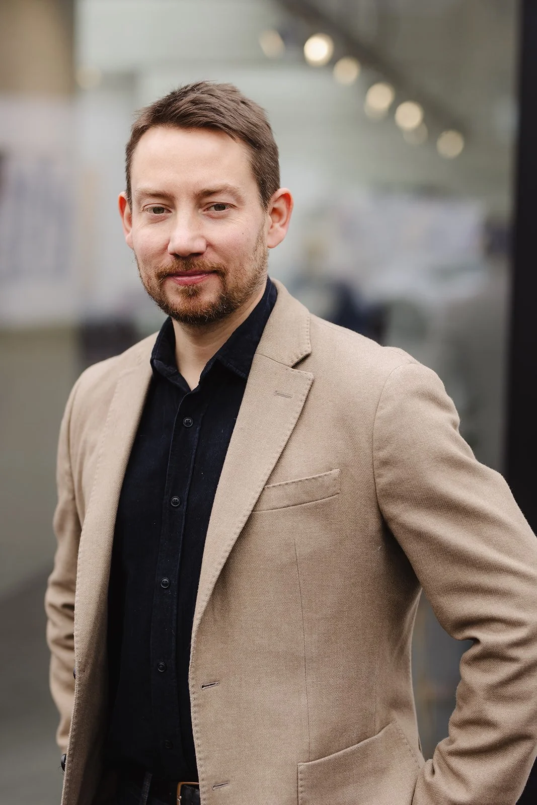 Homme portant un blazer beige et une chemise noire, posé devant une vitre avec des lumières floues en arrière-plan.