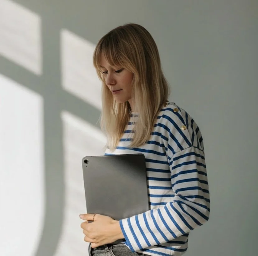 Jeune femme avec cheveux longs et blond foncé, portant un pull à rayures bleues et blanches, tenant une tablette, dans une pièce avec une lumière naturelle.