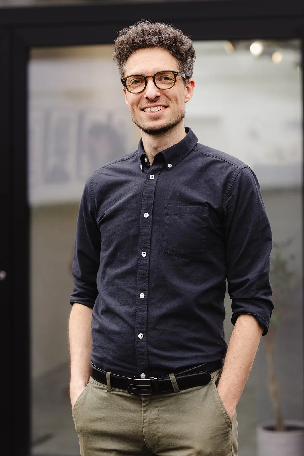 Homme souriant, portant une chemise noire et lunettes, debout en plein air.