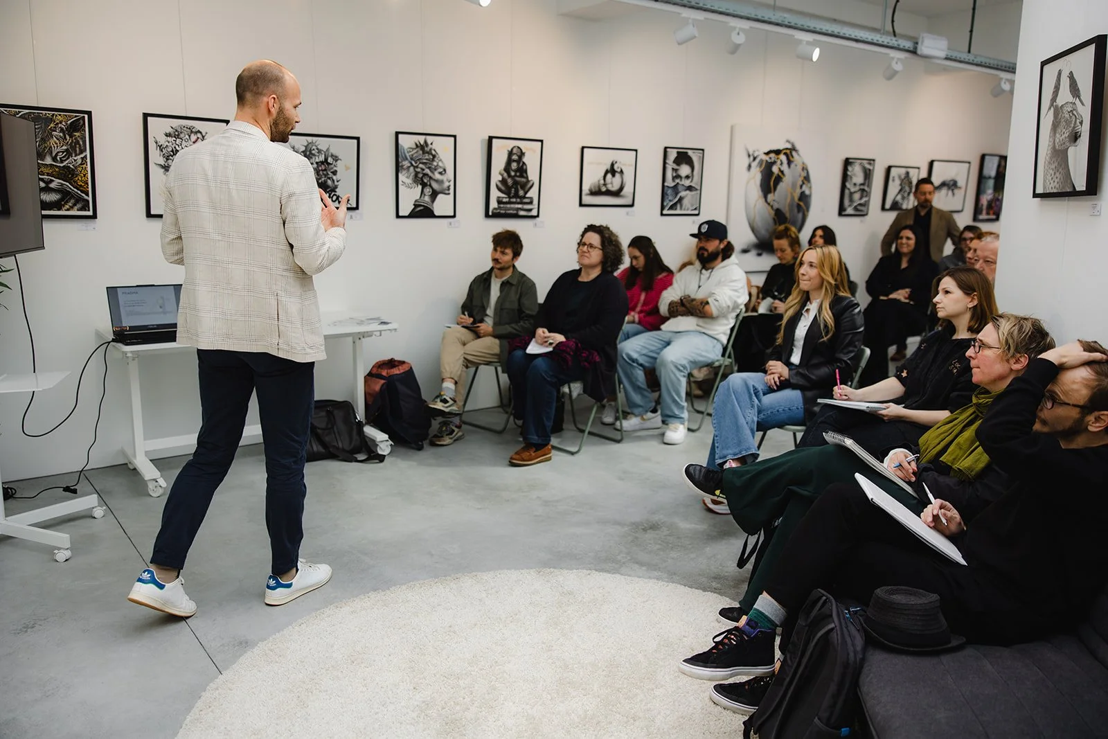 Un homme en costume léger parle à un groupe de personnes lors d'une présentation dans une galerie d'art. Les spectateurs assis prennent des notes et écoutent attentivement, tandis que les œuvres d'art encadrées ornent le mur derrière eux.