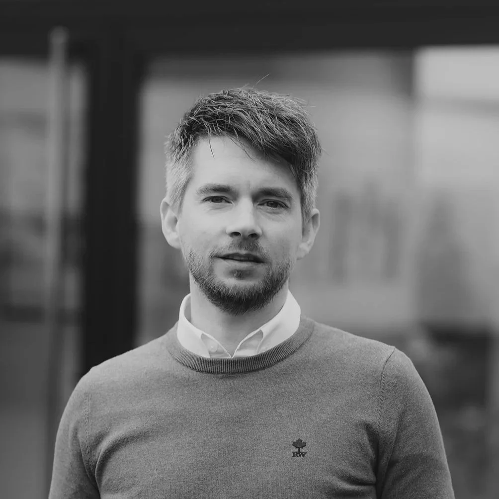 Un portrait en noir et blanc d'un homme jeune avec une barbe et des cheveux courts, portant un pull gris avec un col blanc, debout à l'extérieur, flou en arrière-plan.