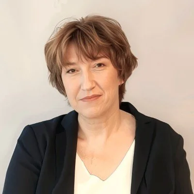 Portrait d'une femme avec coupe de cheveux courte et brun clair, portant une veste noire et un chemisier blanc, souriant légèrement, sur un fond neutre.