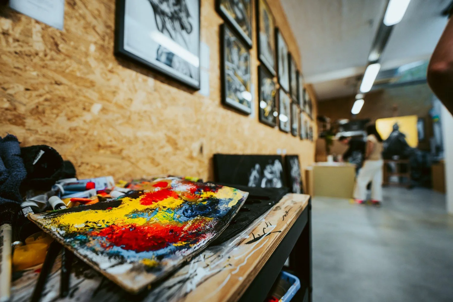 Table de peinture avec palette de couleurs vives, entourée de matériel artistique dans un atelier d'art avec un mur en bois et des œuvres encadrées, et deux personnes en arrière-plan.