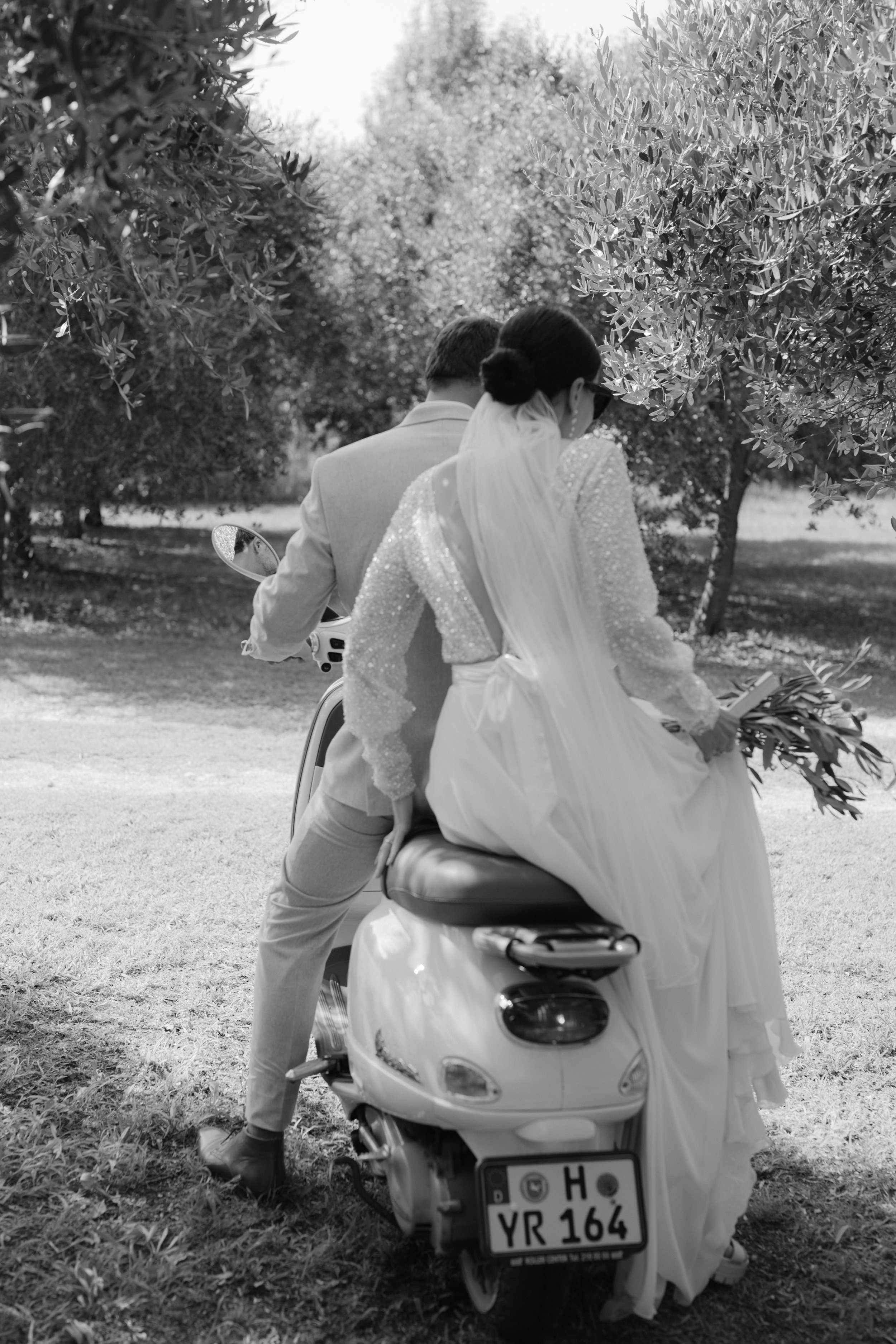 Ein Brautpaar sitzt auf einer Vespa, umgeben von Zypressen und Olivenbäumen am Gardasee.
