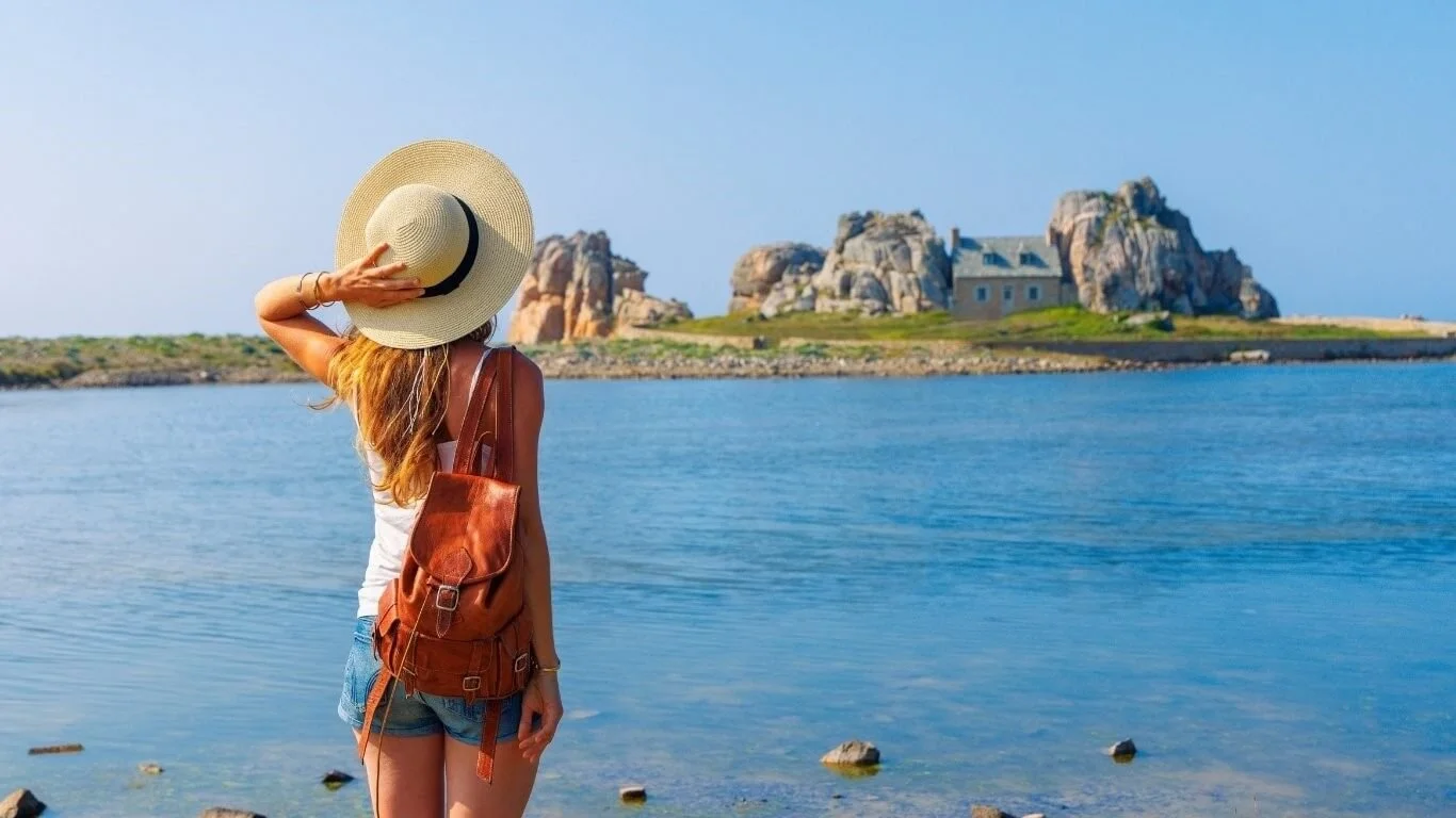 Femme portant un chapeau et un sac à dos regarde la mer avec une île rocheuse en arrière-plan.