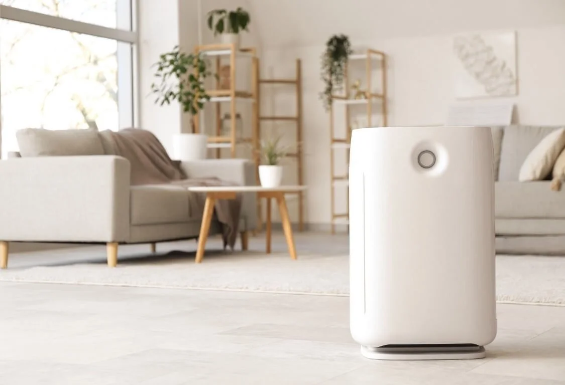 Purificateur d'air blanc dans un salon moderne lumineux avec canapé gris, étagères en bois, et plantes vertes.