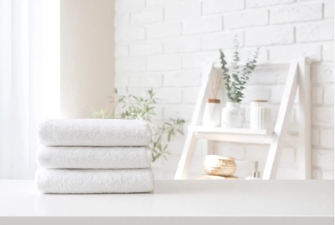 Trois serviettes blanches pliées posées sur une surface blanche, avec un mur blanc en arrière-plan et une étagère en bois portant des vases avec des plantes.