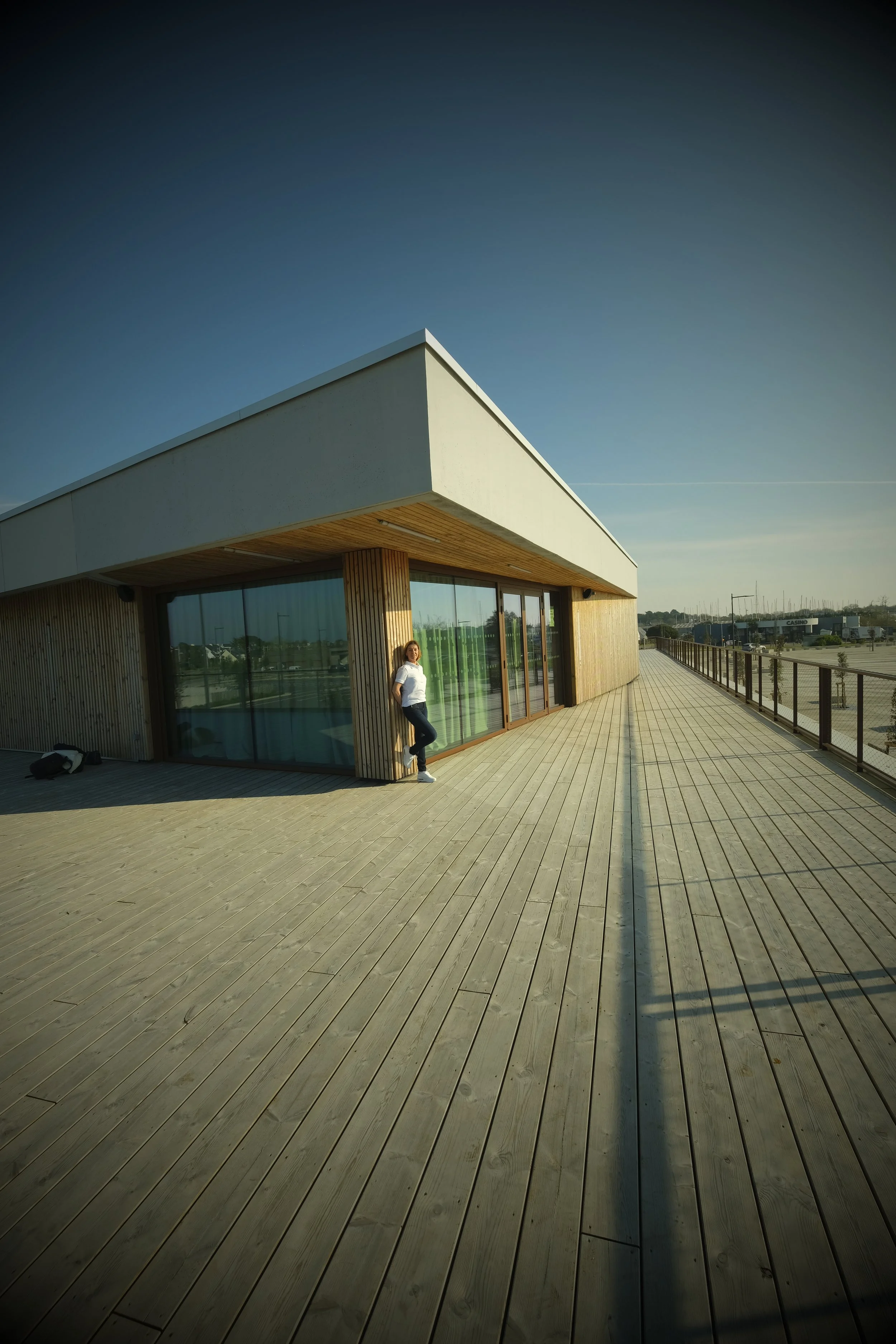 Une femme debout sur une terrasse en bois à côté d'une grande fenêtre d'une construction moderne avec un toit blanc et des murs en bois, sous un ciel clair.