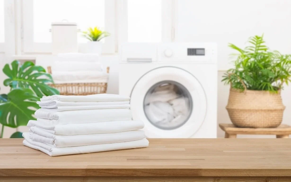 Une pile de linge blanc plié sur une table en bois, avec un lave-linge blanc en arrière-plan, entouré de plantes vertes dans des pots en osier.