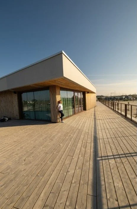 Une personne sur une terrasse en bois avec un bâtiment moderne en arrière-plan, sous un ciel dégagé.