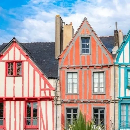 Maisons à colombages colorées avec façades rayées en rouge, orange et bleu, sous un ciel partiellement nuageux.