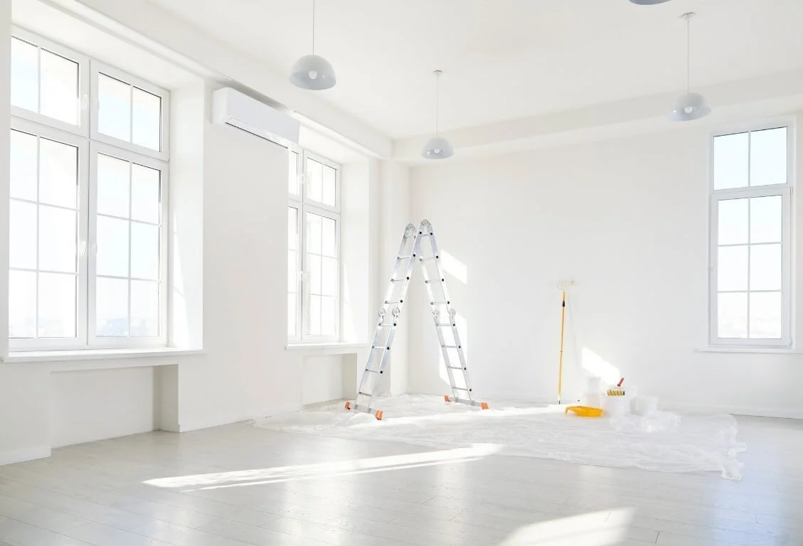 Une pièce lumineuse en cours de rénovation avec des fenêtres, un escabeau en aluminium, un balai, de la peinture et des outils.