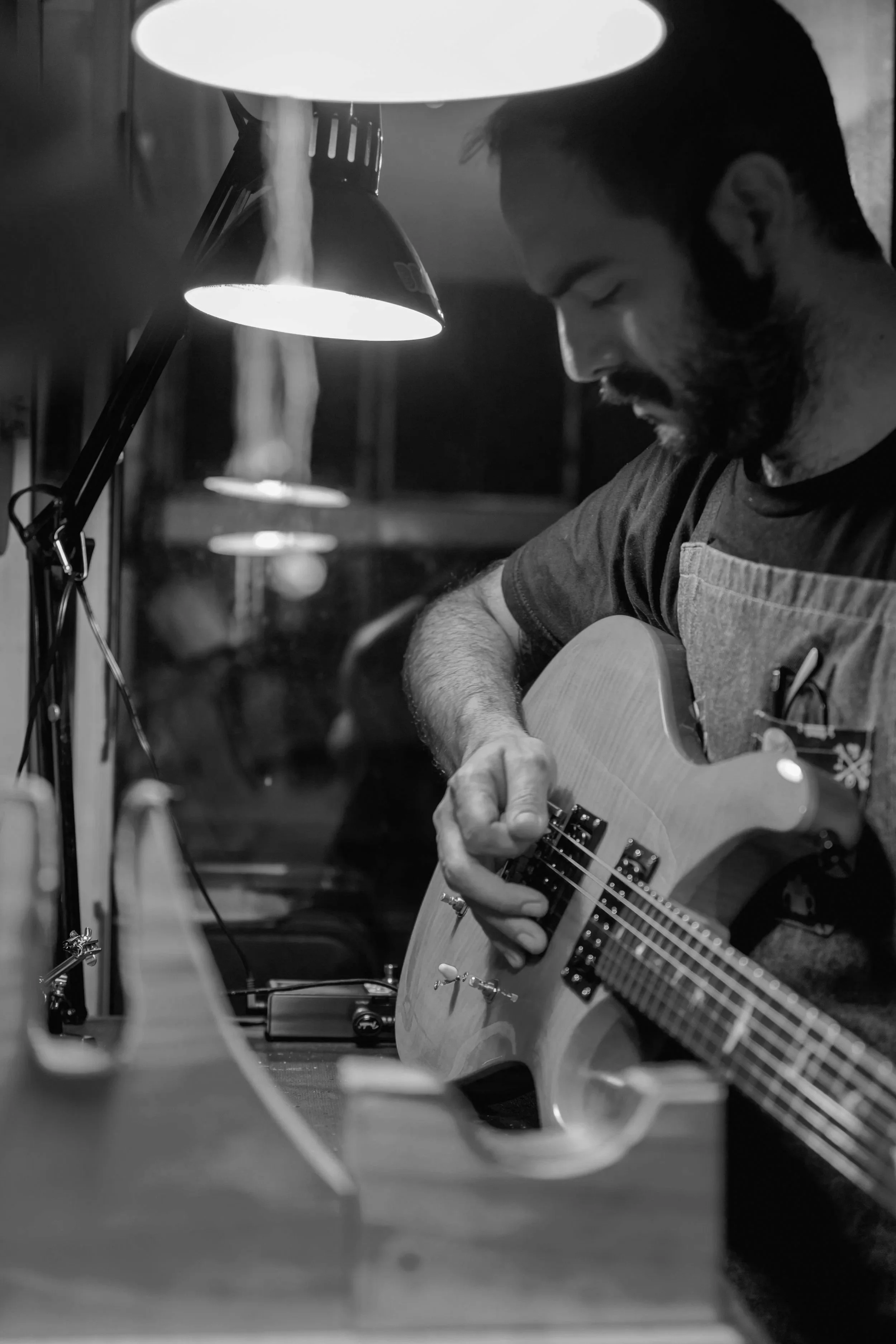 A man playing an acoustic guitar in a dimly lit room, lit by a desk lamp with multiple lamps and a blurred background.