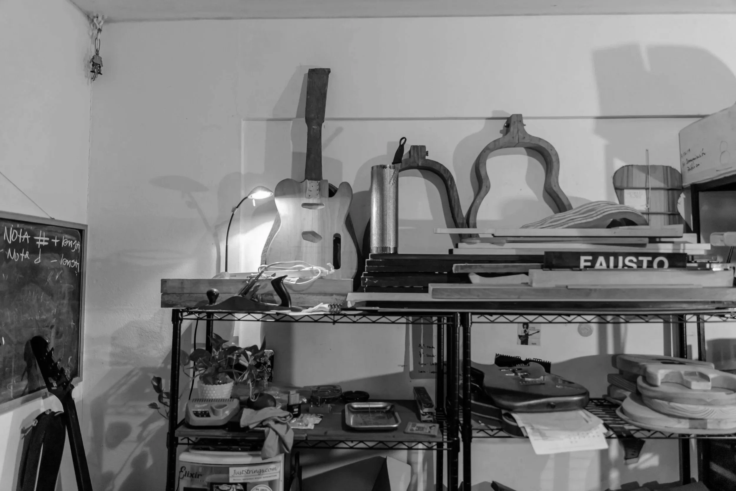 A cluttered storage or workspace area with a metal shelving unit holding various items including books, a plant, a blackboard, and an assortment of tools and objects. The wall behind has a few unidentifiable items hanging.