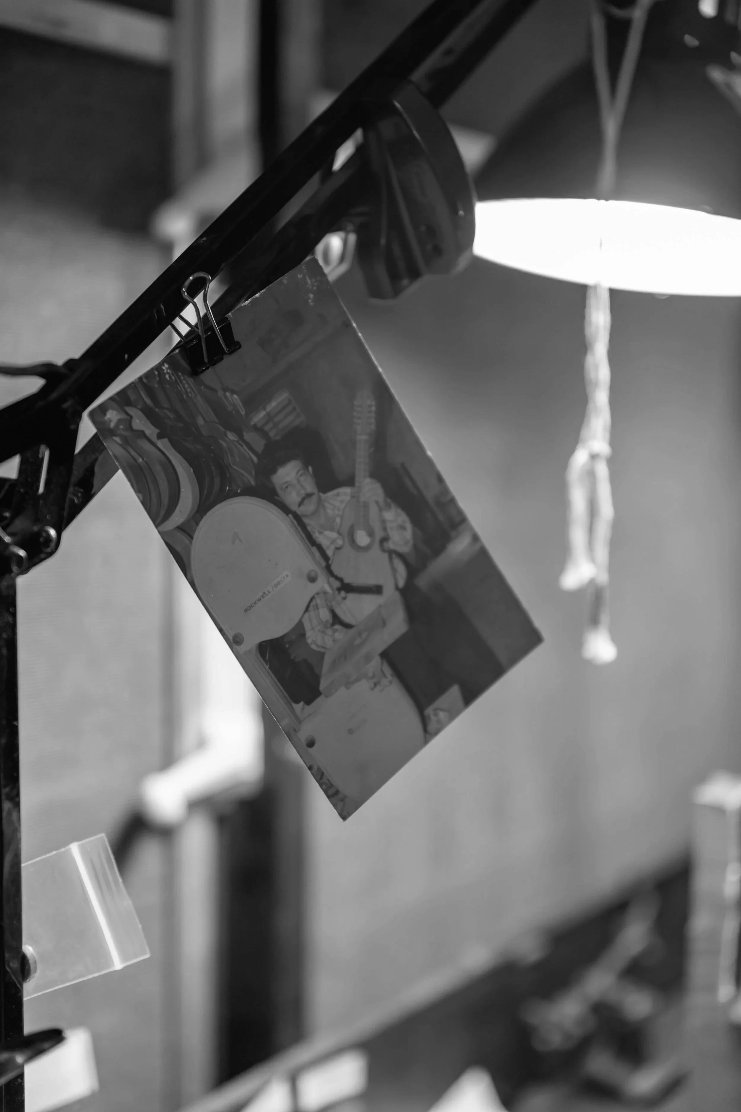 A black and white photo showing a photograph clipped to a display stand. The photograph features a man with dark hair and a mustache, holding a guitar.