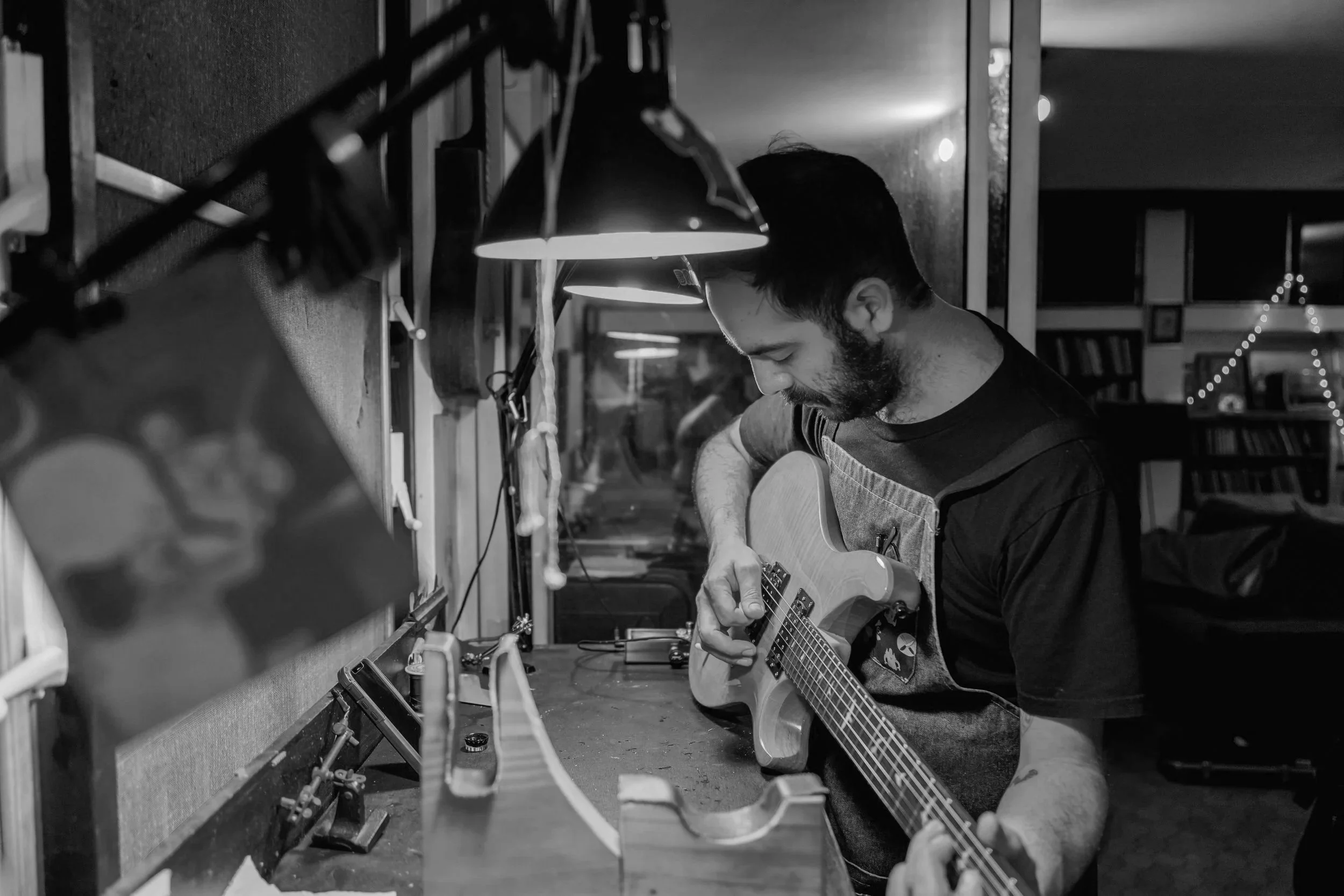 A man with dark hair and a beard, wearing an apron, is sitting on a stool and playing an acoustic guitar in a music or workshop space. There are lamps hanging overhead, illuminating the workspace, and shelves with books and music equipment in the background.