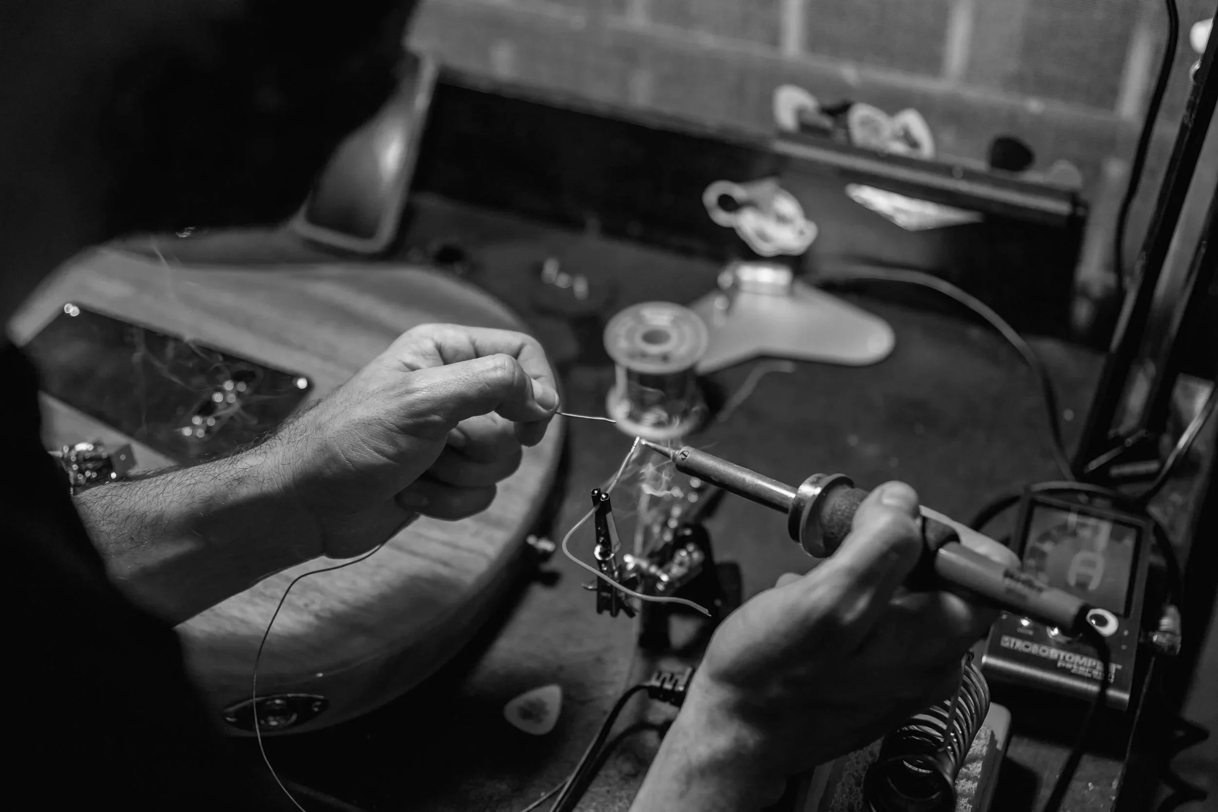 A person working on a soldering project, wiring a component on a workbench in a workshop, with tools and supplies visible in the background.