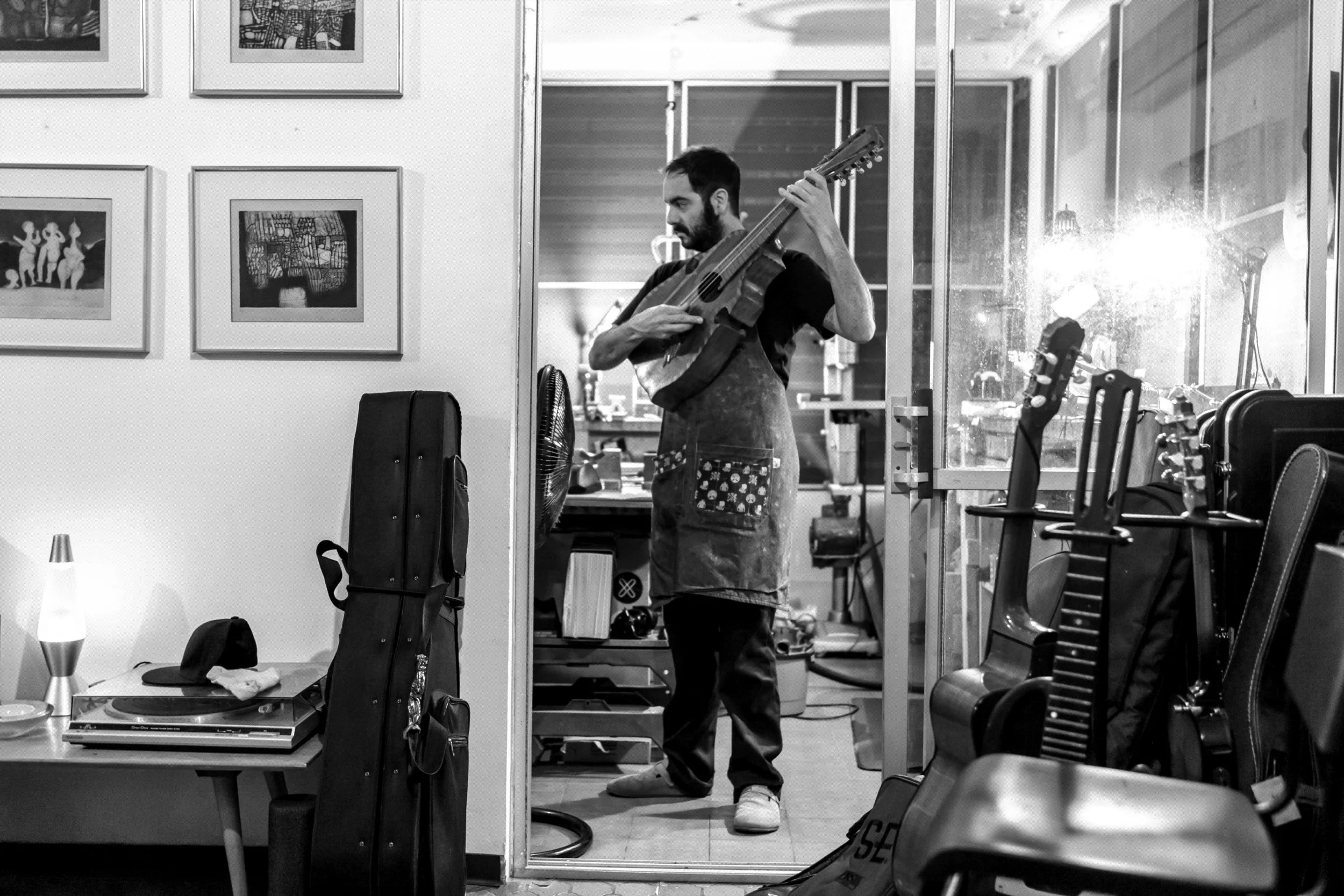 A man playing a guitar in a room with chairs, a table, and wall art, seen through a glass door with reflections.