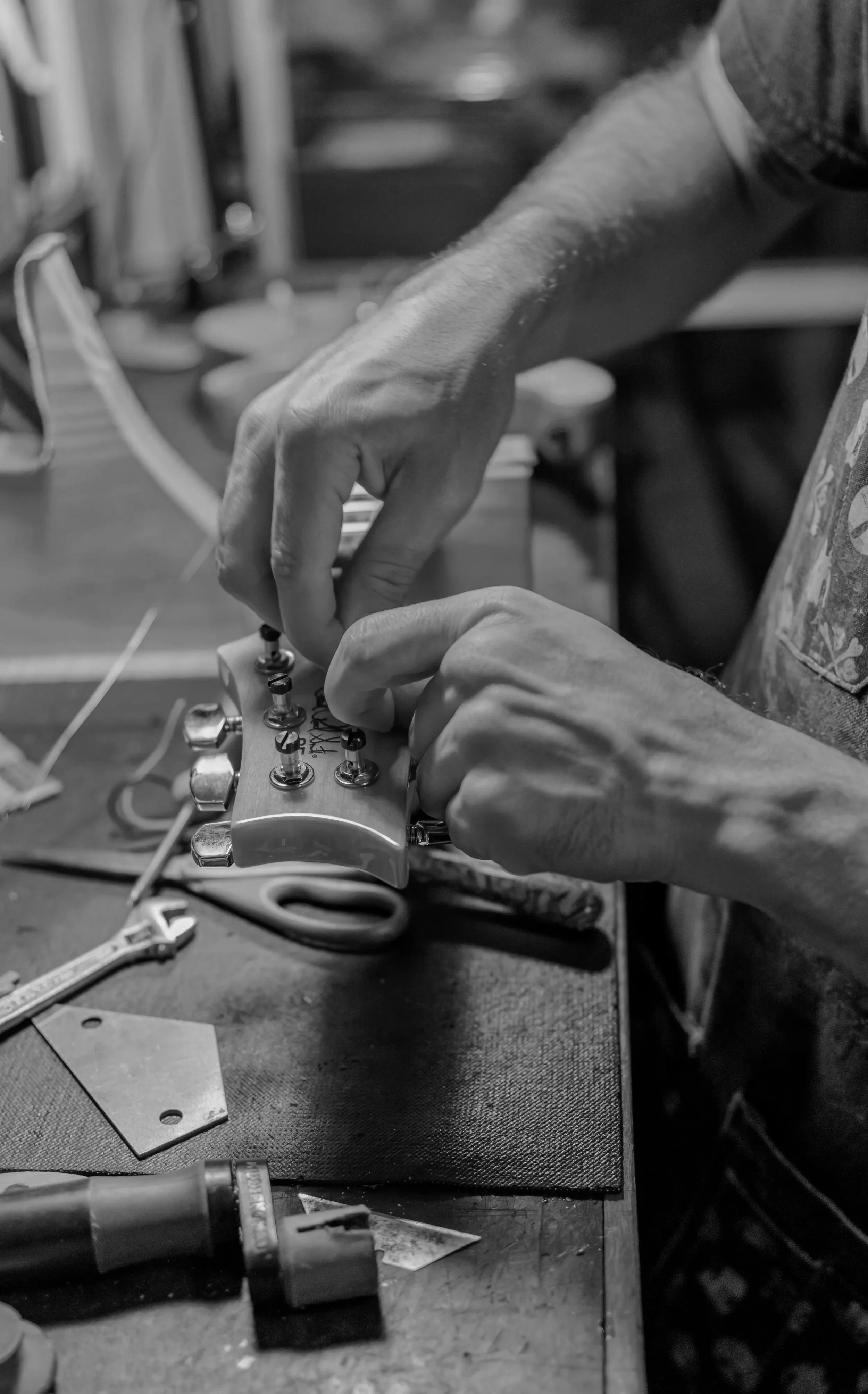 A person adjusts the tuning pegs on a guitar headstock at a workbench, with tools and a soldering iron nearby.