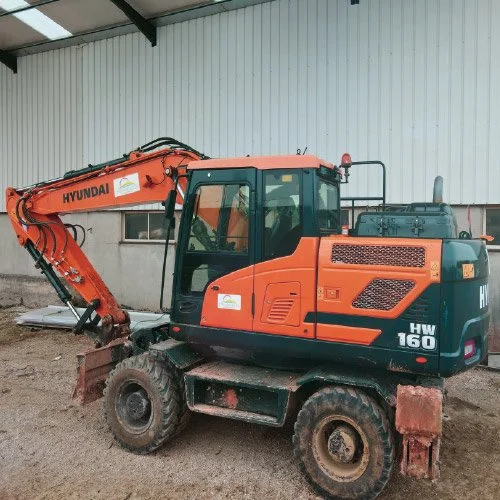 Escavadeira Hyundai HW 160 estacionada em um canteiro de obras, ao lado de uma estrutura de metal.
