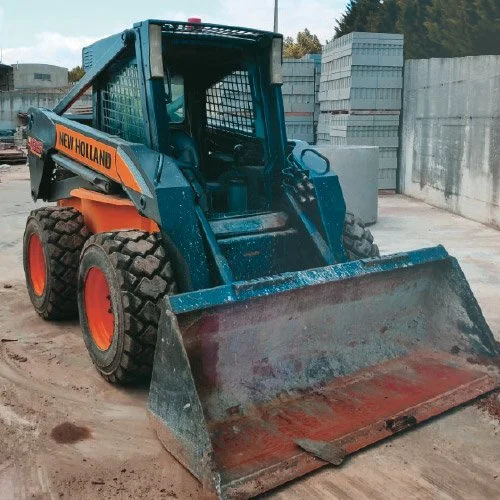 Mini carregadeira azul com rodas grandes, com um balde na frente, marca 'New Holland', em um terreno de construção ao ar livre.
