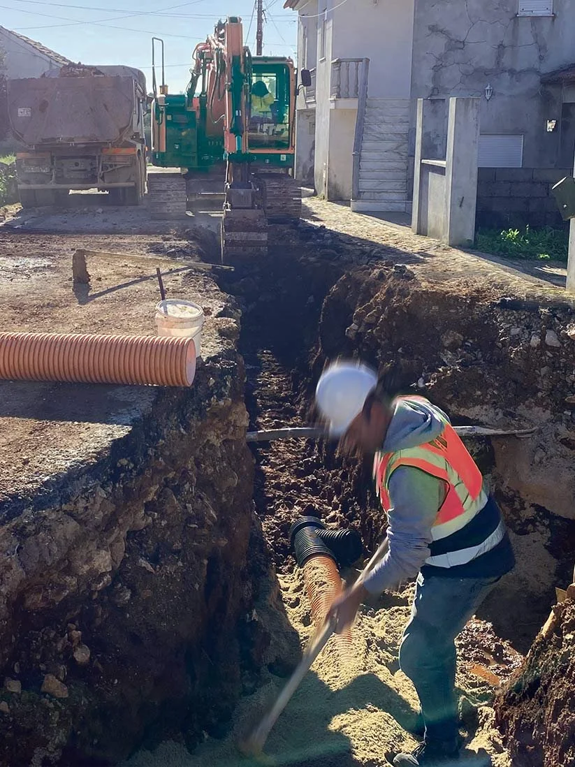 Obra de construção com trabalhador cavando uma vala com ferramenta manual, próximo a equipamentos de escavação e tubos de PVC no local.