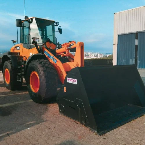 Escavadeira de construção com caçamba volumosa estacionada ao lado de um prédio industrial em um espaço pavimentado.