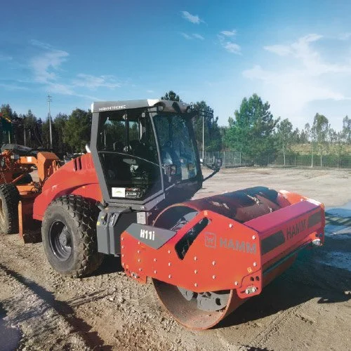 Carro compactador de asfalto vermelho em um canteiro de obras ao ar livre, com grama e árvores ao fundo e céu azul.
