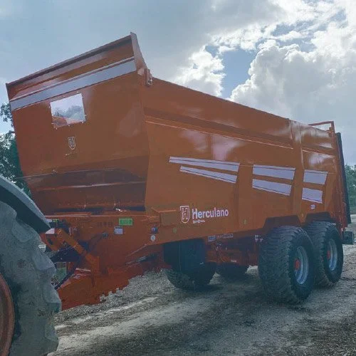 Carro agrícola de cor laranja com marca Hercolano, equipado com duas rodas traseiras e uma dianteira, estacionado em terreno de terra sob céu parcialmente nublado.