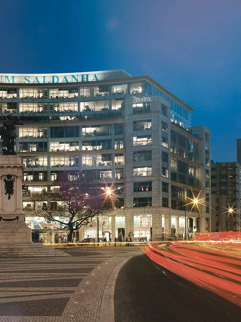 Prédio moderno de vidro em uma cidade à noite, com luzes acesas e passagens de carros com rastros de luz devido à longa exposição.