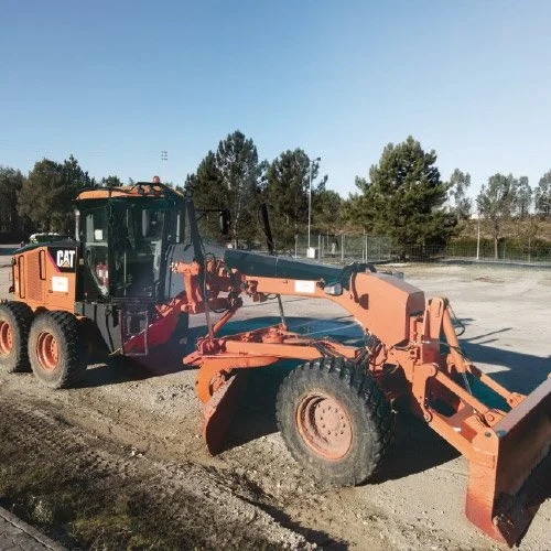 Este é um equipamento de movimento de terra, uma escavadeira carregadeira da marca Caterpillar (CAT), localizada em um local de construção ou manutenção com árvores ao fundo e céu claro.