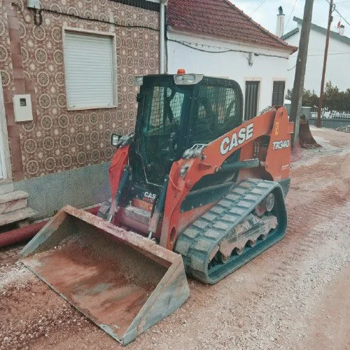 Mini escavadora CASE TR340 em um terreno arenoso ao lado de uma casa com paredes decoradas e uma janela com persiana fechada.