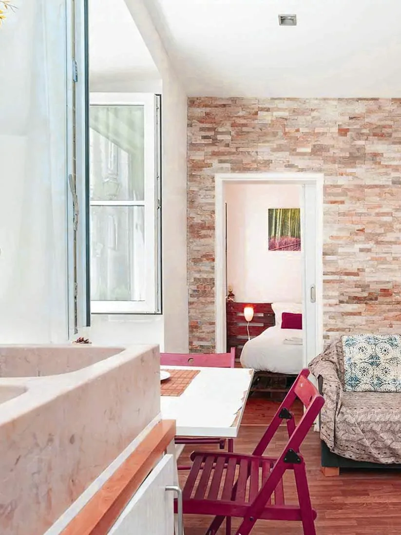 Sala de estar com parede de tijolos expostos, janela aberta, sofá com almofada azul e branca, mesa de jantar com cadeiras vermelhas, vislumbre de quarto com cama, abajur e quadro na parede.