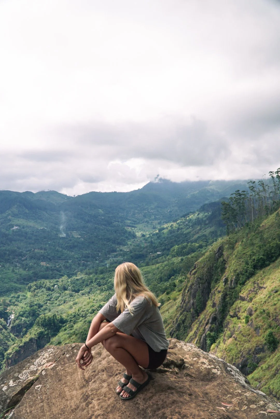 A woman with blonde hair sitting on a large rock, overlooking lush green mountains under an overcast sky.