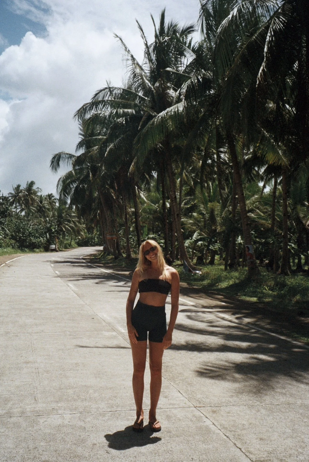 A woman in black summer clothing standing on a sunny street lined with palm trees.