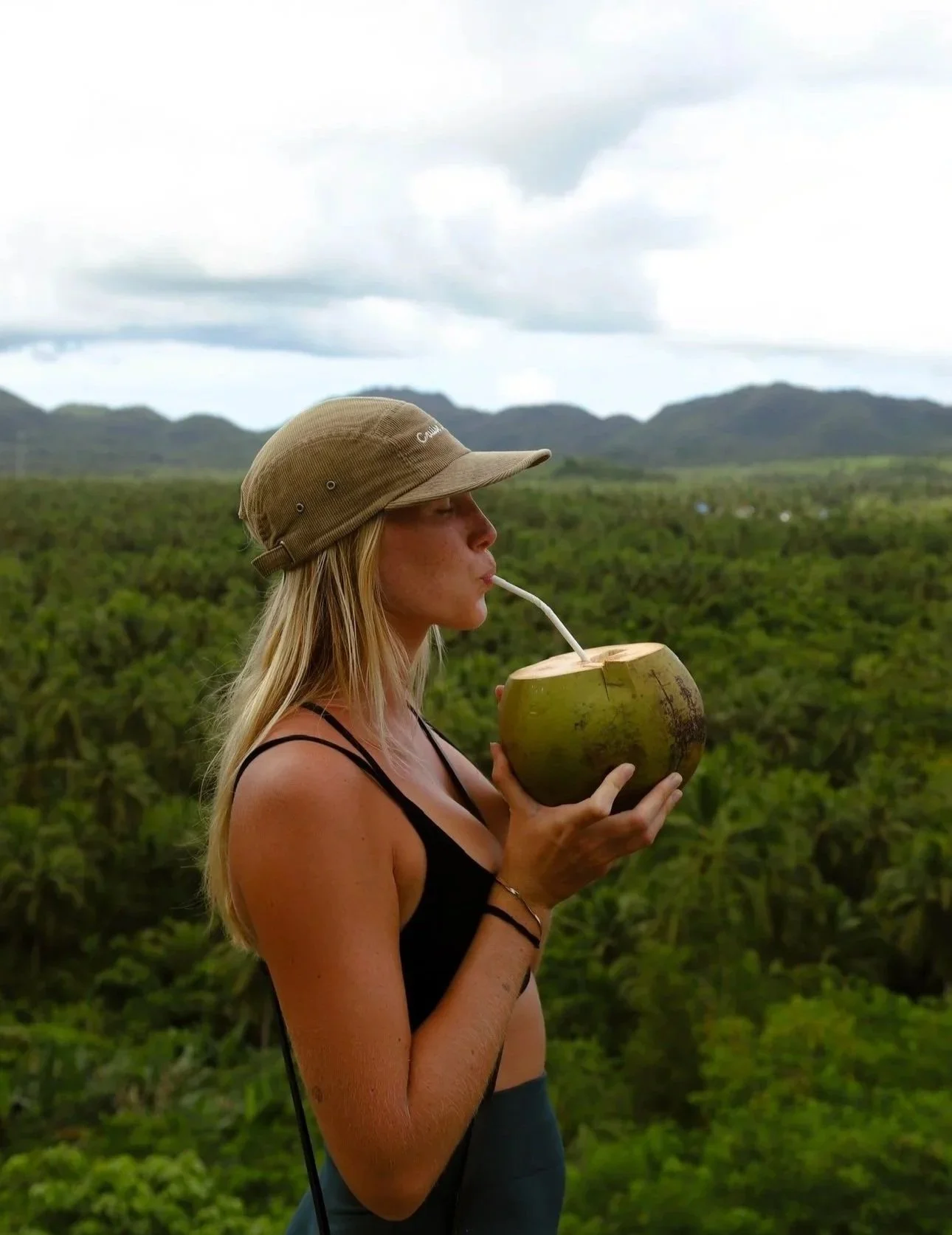 A woman wearing a tan hat and black sports bra drinks from a coconut with a straw against lush green jungle and mountains under a cloudy sky.