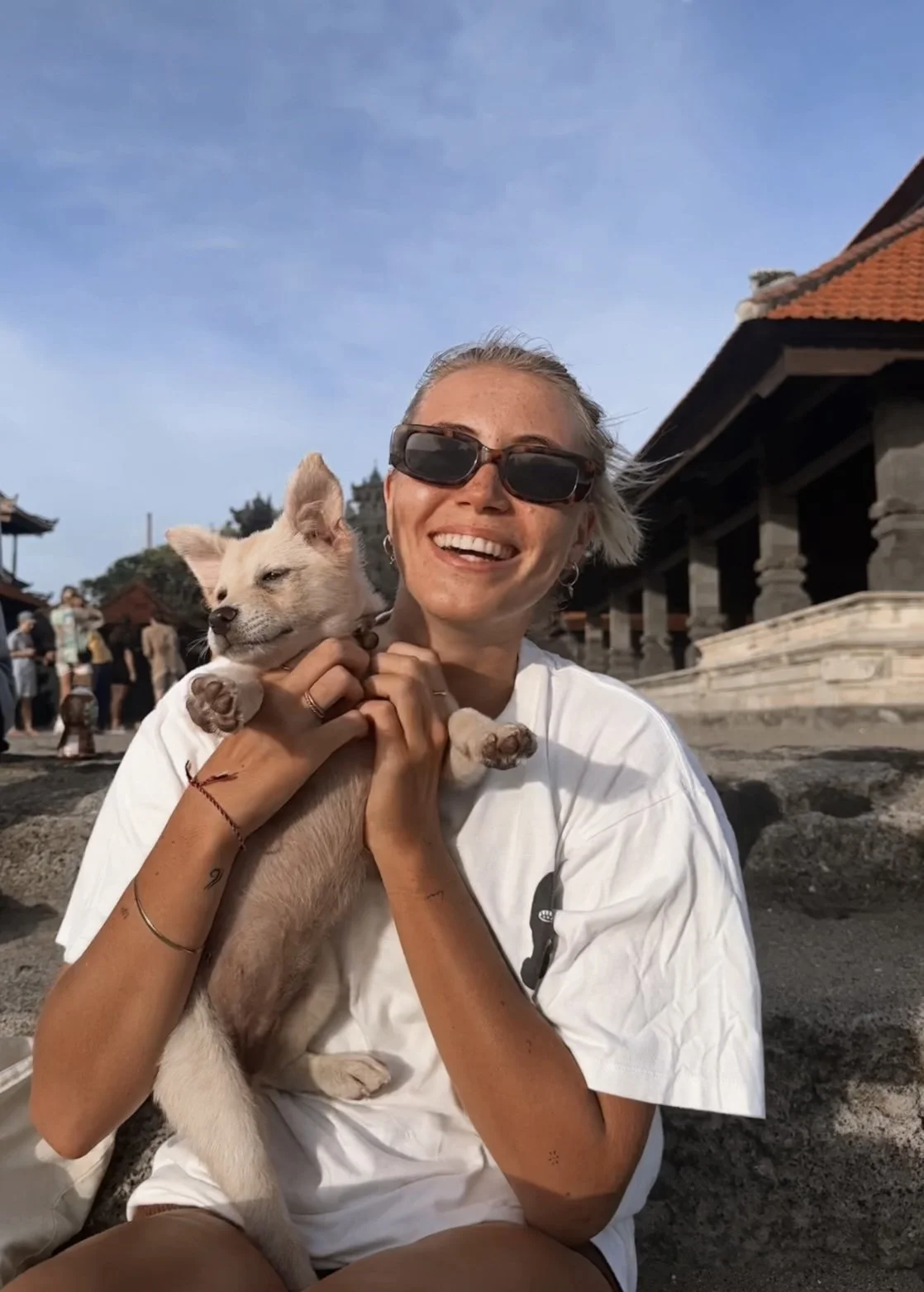 A woman wearing sunglasses smiling and holding a small light-colored puppy with pointed ears outdoors on a sunny day, with traditional buildings and a group of people in the background.