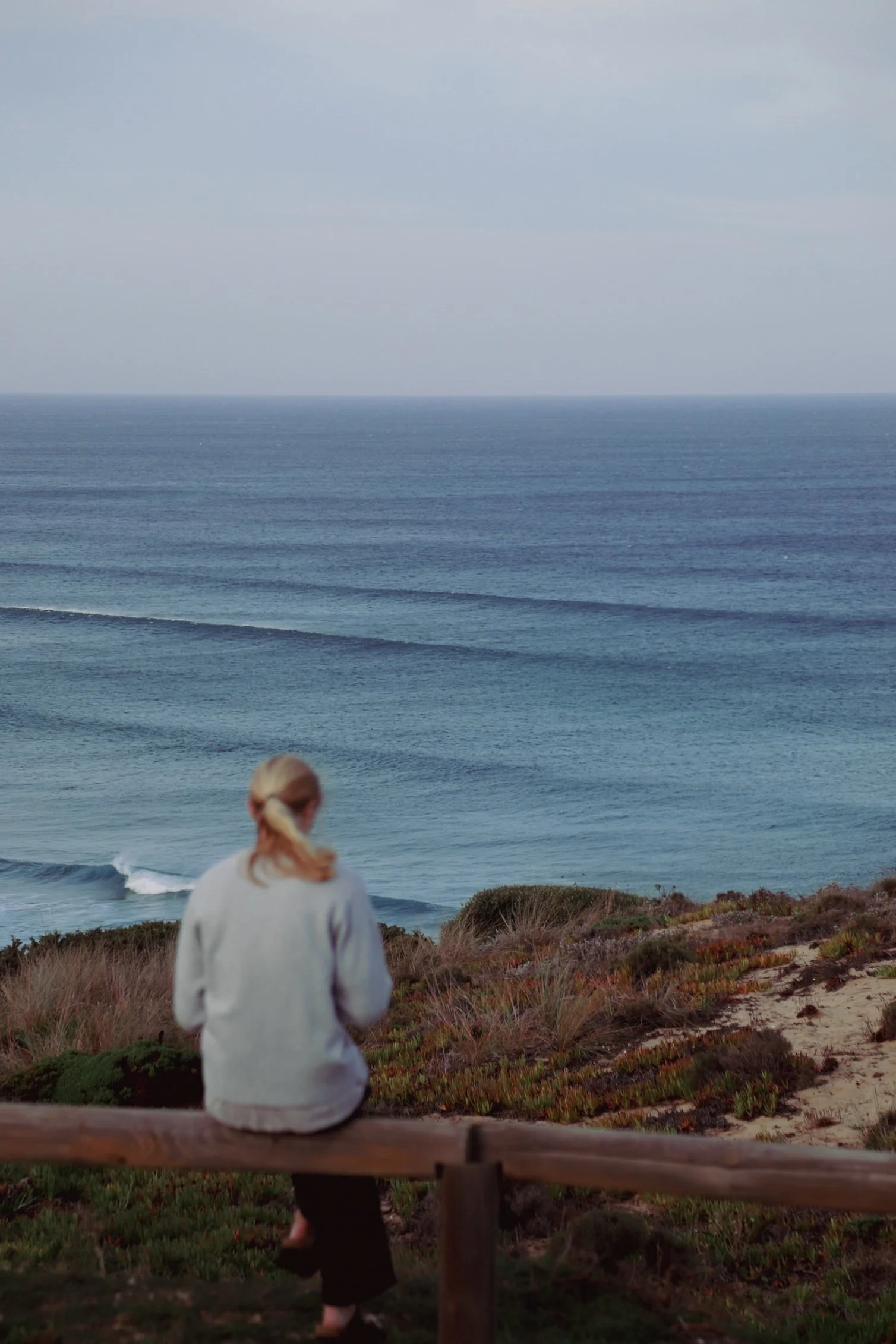 A woman with a ponytail, wearing a light gray jacket, sitting on a wooden barrier overlooking the ocean, with waves in the water and dunes with green and brown vegetation in the foreground.