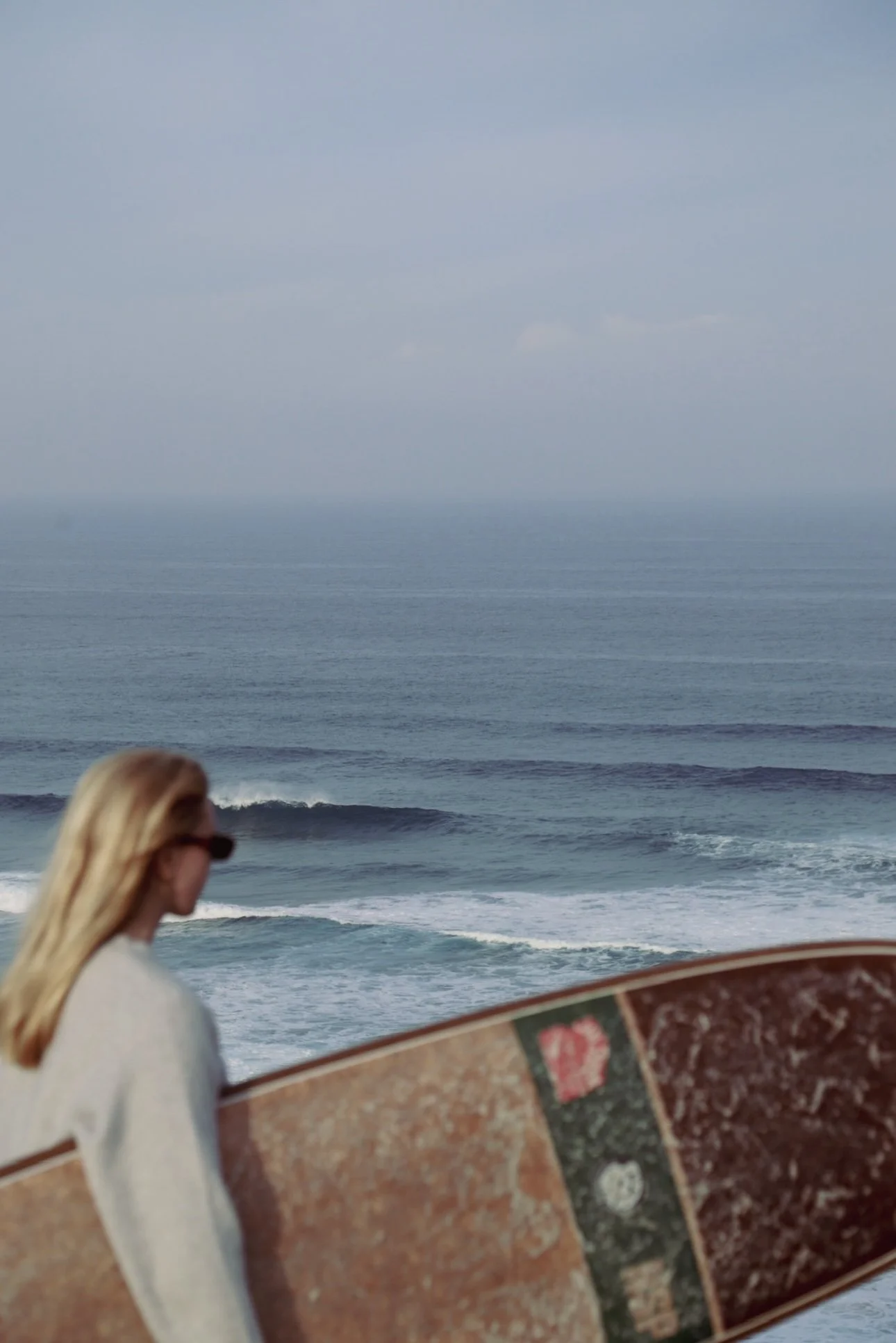 A woman with blonde hair wearing sunglasses, holding a surfboard, standing near the ocean with waves and a cloudy sky.