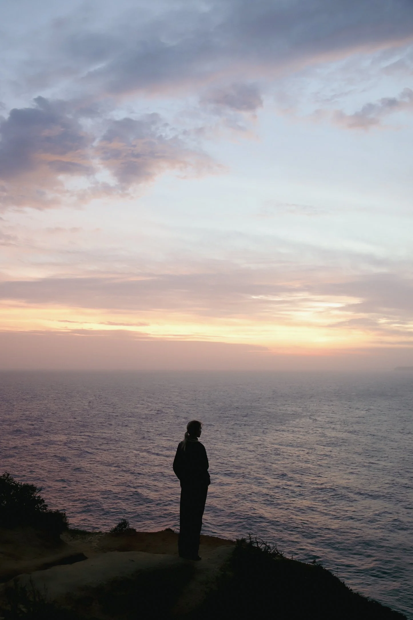 A person stands on a cliff overlooking the ocean at sunset, with clouds in the sky and the water reflecting soft pastel colors.