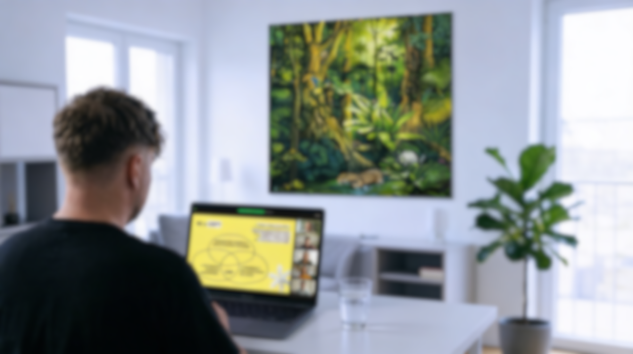 A man working on a laptop with a yellow screen in a bright, modern living room featuring a large abstract forest painting on the wall and a potted plant.
